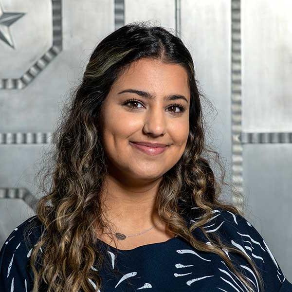 A woman with tanned skin and dark brown hair with light brown highlights poses in front of a silver wall. She wears a navy blue and light blue dress shirt.