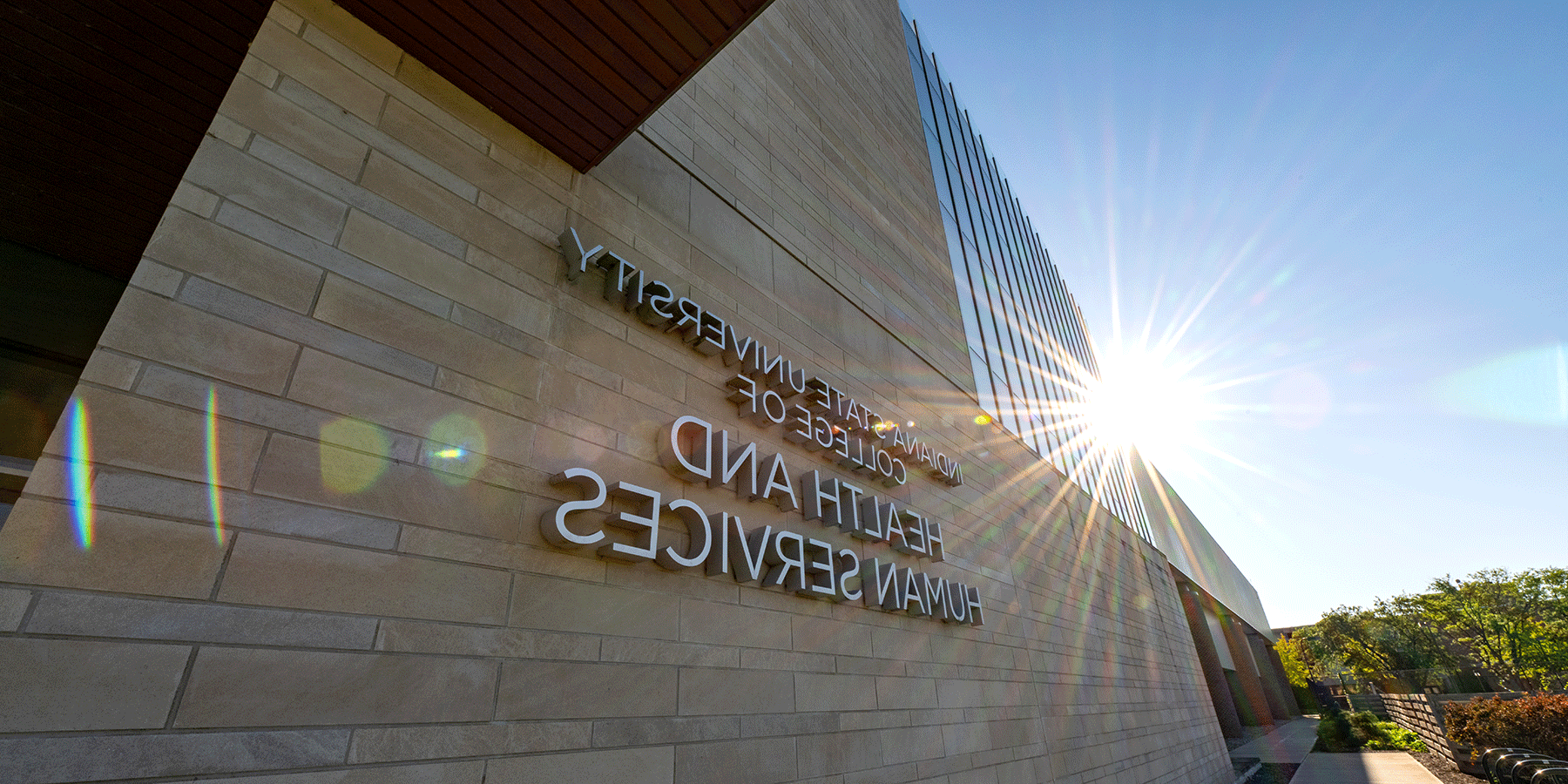 An exterior view of the College of Health and Human Services building, a brick building with glass windows on the left side of the building. “Indiana State University College of Health and Human Services” lettering is on the front side. Green trees are to the left of the building.