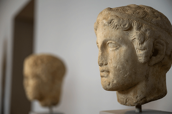 Two portrait busts on display. The sculpture closest to the camera is clearly visible while the other sculpture and background is blurred.