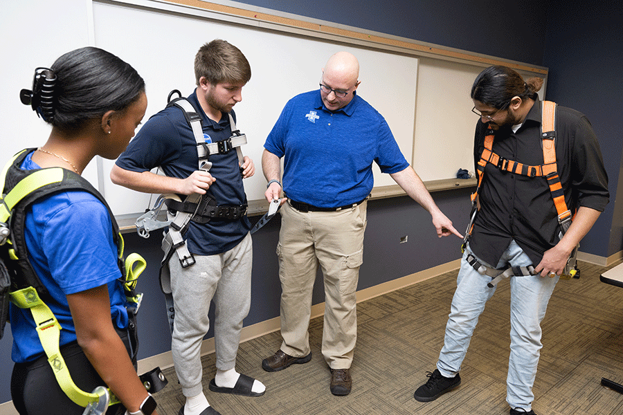 Four individuals are inside a classroom with a whiteboard behind them. On the left is a male student with darker skin. He has long dark brown hair pulled back into a ponytail. He wears a black shirt, denim pants, and an orange safety harness around his shoulders. To his right is a middle-aged white bald male, wearing black glasses, a blue polo shirt, and khaki pants. He is pointing to the student to his left. To his right is a white male student with short brown hair.