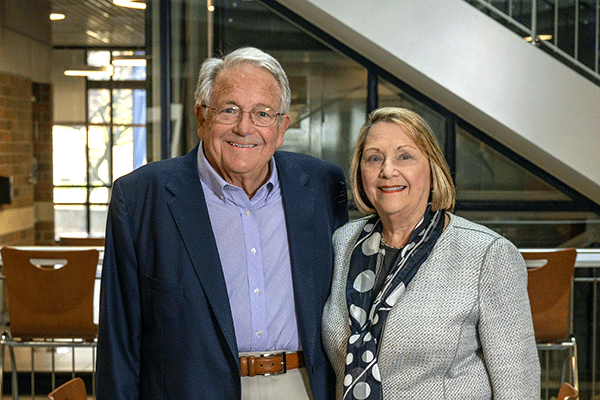 Gloria (left) and Steve (right) Bailey stand smiling in the Bailey College of Engineering & Technology Building. She is wearing a beige business casual suit with a black scarf that has large white circles on it. He is wearing a dark blue sport coat with light blue shirt, beige khakis, and brown belt. The visible interior of the building is dark, with outdoor sunlight visible through windows in the back of the building